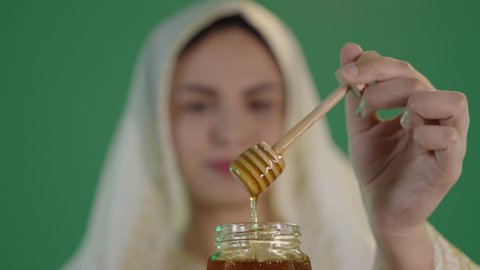 استخراج عسل النحل الطبيعي من المناحل، منتج غذائي محلي وطني صحي في المملكة العربية السعودية،  ارتداء الزي السعودي التقليدي الشعبي , بورتريه لإمرأة عربية خليجية سعودية ترتدي الثوب الابيض المطرز بالذهبي وتضع الشيله على رأسها تحمل بيدها وعاء زجاجي ملئ بالعسل ، كروما ، خلفية خضراء