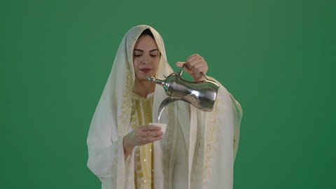 بورتريه لإمرأة عربية خليجية سعودية ترتدي الثوب الابيض المطرز بالذهبي وتضع الشيله على رأسها تقوم بصب فنجان قهوة ساخن، ارتداء الزي السعودي التقليدي الشعبي , حسن الضيافة والاستقبال وإكرام الضيوف، تقديم القهوة العربية في الأعياد والمناسبات ، كروما ، خلفية خضراء