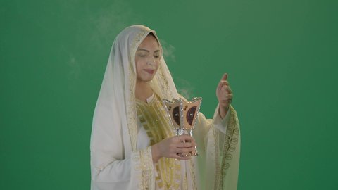 استخدام خشب العود بالأعياد والمناسبات, أصالة العادات والتقاليد الخليجية,ارتداء الزي السعودي التقليدي الشعبي ,بورتريه لإمرأة عربية خليجية سعودية ترتدي الثوب الابيض المطرز بالذهبي وتضع الشيله على رأسها  تمسك المبخرة التراثية المزخرفة بيدها و تنظر إلى الكاميرا بإيماءات السعادة و السرور, مفهوم التعطر بالبخور, كروما ، خلفية خضراء