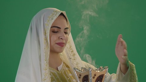 أصالة العادات والتقاليد الخليجية, استخدام خشب العود بالأعياد والمناسبات, ارتداء الزي السعودي التقليدي الشعبي ,بورتريه لإمرأة عربية خليجية سعودية ترتدي الثوب الابيض المطرز بالذهبي وتضع الشيله على رأسها  تمسك المبخرة التراثية المزخرفة بيدها و تنظر إلى الكاميرا بإيماءات السعادة و السرور, مفهوم التعطر بالبخور, كروما ، خلفية خضراء