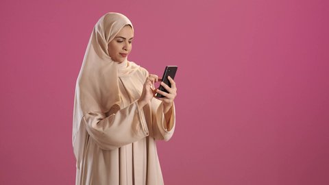 Browsing applications and social media programs, exchanging conversations with family and friends, a portrait of a Saudi Gulf Arab woman wearing an abaya and hijab holding a mobile phone in her hand, using modern advanced devices and technologies, red background.