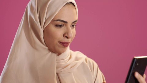 استخدام الأجهزة والتقنيات الحديثة المتطورة، تصفح التطبيقات وبرامج التواصل الاجتماعي، تبادل الأحاديث مع الأهل والأصدقاء، بورتريه لامرأة عربية خليجية سعودية ترتدي العباءة والحجاب تمسك بيدها الجوال ، خلفية حمراء