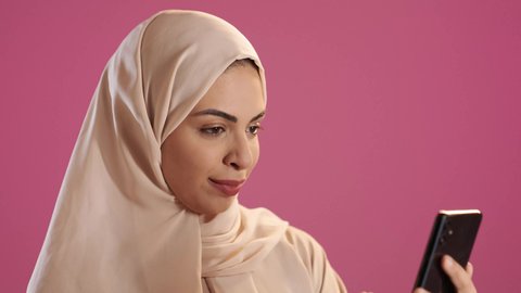 Browsing applications and social media programs, exchanging conversations with family and friends, a portrait of a Saudi Gulf Arab woman wearing an abaya and hijab holding a mobile phone in her hand, using modern advanced devices and technologies, red background.