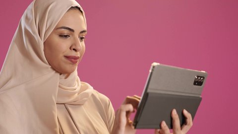 الدردشة و التواصل الاجتماعي, إجراء مكالمة فيديو عبر التابلت, ايماءات السرور والبهجة, بورتريه لامرأة عربية خليجية سعودية ترتدي العباءة والحجاب تنظر الى الكاميرا بايماءات السعادة , تقنيات العمل عن بعد ، خلفية حمراء