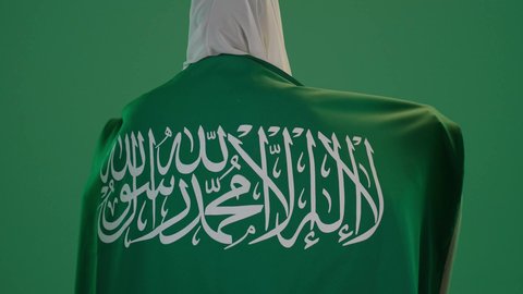 ذكرى يوم بدينا 1727 م، تأسيس الدولة السعودية الأولى 22 فبراير، ارتداء الزي السعودي التقليدي الشعبي , بورتريه من الخلف لإمرأة عربية خليجية سعودية ترتدي الثوب الابيض المطرز بالذهبي وتضع الشيله على رأسها تمسك راية وشعار يوم التأسيس بتعابير الفخر والاعتزاز, كروما ، خلفية خضراء