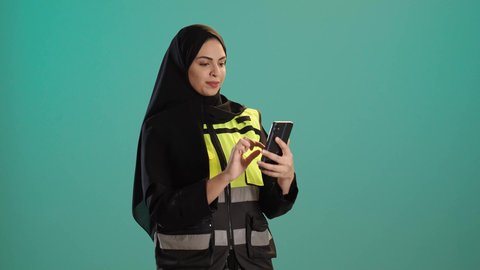 العمل في القطاع الهندسي, دمج التكنولوجيا الحديثة بالمجال الهندسي، وظائف ومهن نسائية,بورتريه لمهندسة عربية خليجية امارتية ترتدي خوذة وسترة الحماية تقوم باجراء مكالمة فيديو، شابة سعودية تحمل بيدها الهاتف المحمول، كروما ، خلفية خضراء