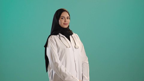 The concept of medicine and healthcare, working in the health sector in the Kingdom, a portrait of an Arab Gulf Saudi female doctor wearing an abaya and hijab with a white coat standing upright, looking with gestures of happiness and joy, female professions and jobs, providing services to patients with efficiency, chroma, green background.