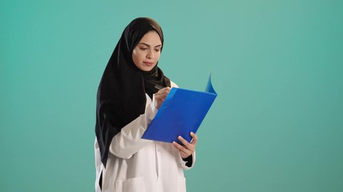 Providing healthcare to patients, writing medical reports, a portrait of an Arab Gulf Saudi female doctor wearing an abaya and hijab with a white coat, wearing a stethoscope around her neck, holding patient papers and files in her hand, working in the healthcare sector in the Kingdom of Saudi Arabia, chroma, green background.