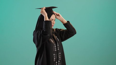 The concept of diligence and excellence, the feeling of pride and self-confidence, celebrating the completion of university studies, a portrait of a young Arab Gulf Saudi woman wearing an abaya and hijab putting on a graduation cap, chroma, green background.