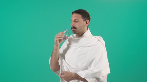 Increasing fluid intake to keep the body from dehydration, performing the rituals of Hajj and Umrah, a close-up portrait of an Arab Saudi Gulf man wearing the Ihram attire drinking fresh water, quenching his thirst with Zamzam water, green background, chroma.