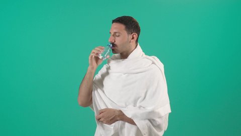 أداء مناسك الحج والعمرة، الاكثار من السوائل للحفاظ على الجسم من الجفاف، بورتريه مقرب لرجل عربي سعودي خليجي يرتدي زي الاحرام يقوم بشرب المياه العذب، الارتواء بماء زمزم، خلفية خضراء ، كروما