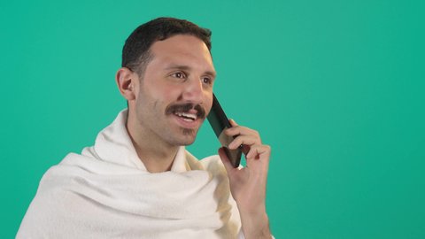 Chatting with family and friends, using a modern technological device, an Arab Gulf Saudi man wearing the Ihram garment holding a mobile phone, integrating technology and technology into daily life, browsing social media.