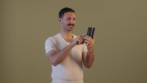 Using a modern technical device, an Arab Gulf Saudi man wearing pants and a white t-shirt is holding a mobile phone in his hand, integrating technology into daily life, browsing social media.