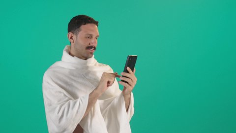 Making a phone call, chatting with family and friends, using modern technology, an Arab Gulf Saudi man wearing the Ihram garment holding a mobile phone, integrating technology and tech into daily life, browsing social media.