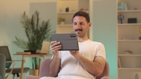 Using tablet applications and programs, chatting with family and friends, integrating technology and modern techniques into daily life, an Arab Gulf Saudi man wearing pants and a white tank top holding a tablet in his hand, browsing social media.