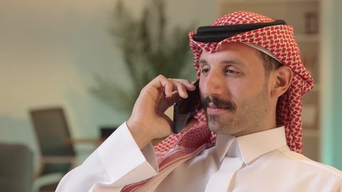 Making a phone call, chatting with family and friends, using modern technology, an Arab Gulf Saudi man wearing traditional attire and a ghutrah holding a mobile phone, integrating technology into daily life, browsing social media.