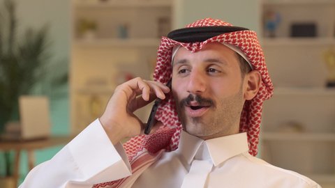 Making a phone call, chatting with family and friends, using modern technology, an Arab Gulf Saudi man wearing traditional attire and a ghutrah holding a mobile phone, integrating technology into daily life, browsing social media.