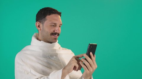رجل عربي خليجي سعودي يرتدي لباس الاحرام يمسك بيده الهاتف المحمول، دمج التقنية والتكنولوجيا بالحياة اليومية، تصفح مواقع التواصل الاجتماعي،اجراء مكالمة هاتفية ، الدردشة مع الأهل والأصدقاء، استخدام جهاز تقني حديث