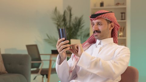 Making a phone call, chatting with family and friends, using modern technology, an Arab Gulf Saudi man wearing traditional attire and a ghutrah holding a mobile phone, integrating technology into daily life, browsing social media.
