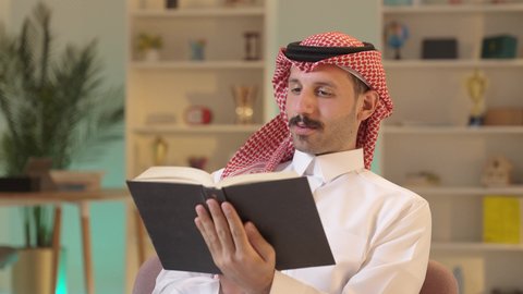 رجل عربي خليجي سعودي يرتدي الزى التقليدي والشماغ يجلس على كرسي مريح يقوم بقراءة القصص ، الأنشطة الترفيهية المفيدة, الاستمتاع بممارسة هواية المطالعة،الروايات والكتب التثقيفية