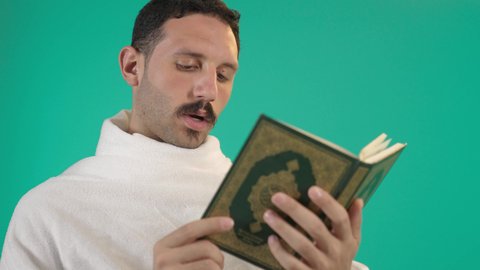 Reflecting on the Quranic verses and contemplating them, pondering the chapters of the Holy Quran, the concept of worship, obedience, and drawing closer to God during the blessed month of Ramadan, an Arab Gulf Saudi man wearing the Ihram clothing reading from the Holy Quran, green background, chroma.