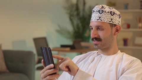 رجل عربي خليجي عماني يرتدي الدشداشة والكمة يمسك بيده الهاتف المحمول، دمج التقنية والتكنولوجيا بالحياة اليومية، الدردشة مع الأهل والأصدقاء، استخدام جهاز تقني حديث،تصفح مواقع التواصل الاجتماعي