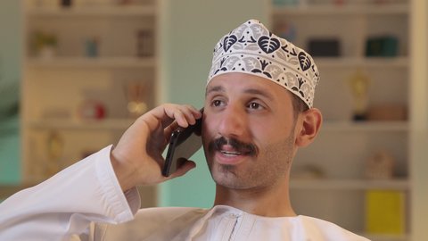 اجراء مكالمة هاتفية ، رجل عربي خليجي عماني يرتدي الدشداشة والكمة يمسك بيده الهاتف المحمول، دمج التقنية والتكنولوجيا بالحياة اليومية،الدردشة مع الأهل والأصدقاء، استخدام جهاز تقني حديث