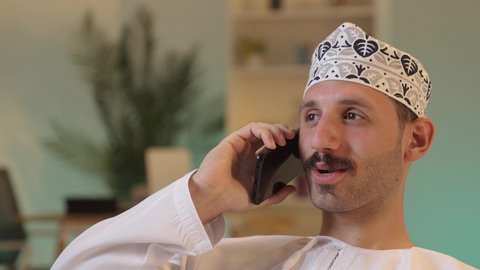 Chatting with family and friends, using a modern technological device, an Arab Gulf Omani man wearing a dishdasha and a kumma holding a mobile phone, integrating technology into daily life, browsing social media.
