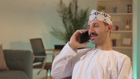 استخدام جهاز تقني حديث، الدردشة مع الأهل والأصدقاء، رجل عربي خليجي عماني يرتدي الدشداشة والكمة يمسك بيده الهاتف المحمول، دمج التقنية والتكنولوجيا بالحياة اليومية، تصفح مواقع التواصل الاجتماعي