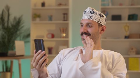 الدردشة مع الأهل والأصدقاء، استخدام جهاز تقني حديث، رجل عربي خليجي عماني يرتدي الدشداشة والكمة يمسك بيده الهاتف المحمول، دمج التقنية والتكنولوجيا بالحياة اليومية، تصفح مواقع التواصل الاجتماعي