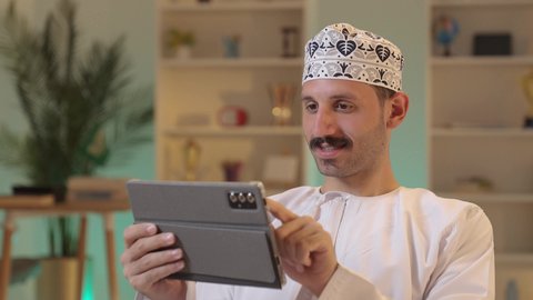 رجل عربي خليجي عماني يرتدي الدشداشة والكمة يحمل بيده جهاز التابلت، استخدام تطبيقات وبرامج الأجهزة اللوحية، الدردشة مع الأهل والأصدقاء، دمج التكنولوجيا والتقنيات الحديثة فى الحياه اليوميه ،تصفح مواقع التواصل الاجتماعي