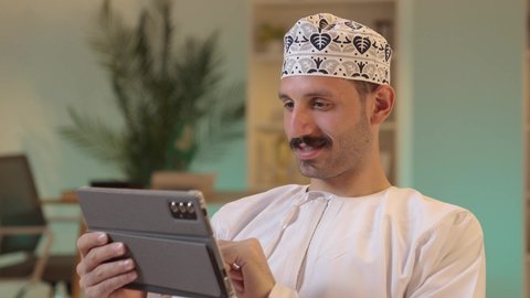 دمج التكنولوجيا والتقنيات الحديثة فى الحياه اليوميه ، استخدام تطبيقات وبرامج الأجهزة اللوحية، الدردشة مع الأهل والأصدقاء، رجل عربي خليجي عماني يرتدي الدشداشة والكمة يحمل بيده جهاز التابلت، تصفح مواقع التواصل الاجتماعي