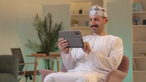 استخدام تطبيقات وبرامج الأجهزة اللوحية، الدردشة مع الأهل والأصدقاء، دمج التكنولوجيا والتقنيات الحديثة فى الحياه اليوميه ، رجل عربي خليجي عماني يرتدي الدشداشة والكمة يحمل بيده جهاز التابلت، تصفح مواقع التواصل الاجتماعي
