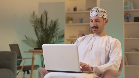 Browsing social media, managing and organizing work affairs, an Arab Gulf Omani man wearing a dishdasha and kumma is working on a laptop with focused gestures, using advanced modern technologies.