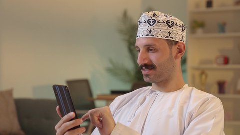 Chatting with family and friends, using a modern technological device, an Arab Gulf Omani man wearing a dishdasha and a kumma holding a mobile phone, integrating technology into daily life, browsing social media.