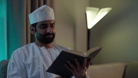 الاستمتاع بممارسة هواية المطالعة، الأنشطة الترفيهية المفيدة,رجل عربي خليجي عماني يرتدي الدشداشة والكمه العمانية يجلس فى غرفه المعيشة يقوم بقراءة القصص ، الروايات والكتب التثقيفية