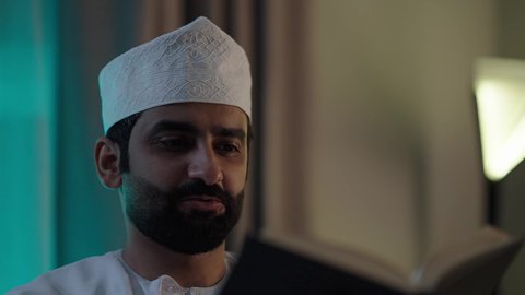 Useful recreational activities, enjoying the hobby of reading, an Arab Gulf Omani man wearing a dishdasha and an Omani kumma sits in the living room reading stories, novels, and educational books.