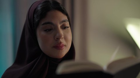 A Gulf Arab Omani woman wearing an abaya and hijab sits in the living room reading stories, novels, and educational books, engaging in useful recreational activities, and enjoying her hobby of reading.