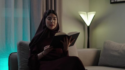 Enjoying the hobby of reading, useful recreational activities, an Arab Gulf Omani woman wearing an abaya and hijab sits in the living room reading stories, novels, and educational books.