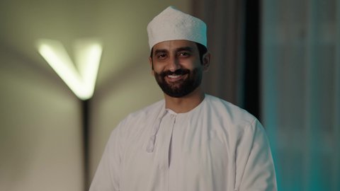 مفهوم الرجولة والوقار، الوقوف بيدين متقاطعتين بإيماءات الثقة بالنفس، الاهتمام بالمظهر الخارجي، بورتريه لرجل عربي خليجي عماني يرتدي الدشداشة التقليدية والكمه العمانية ينظر بايماءات السرور والسعادة