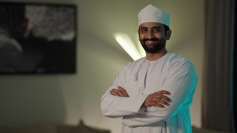 Attention to appearance, standing with arms crossed in gestures of self-confidence, the concept of masculinity and dignity, a portrait of an Arab Gulf Omani man wearing the traditional dishdasha and Omani kumma, looking with gestures of joy and happiness.