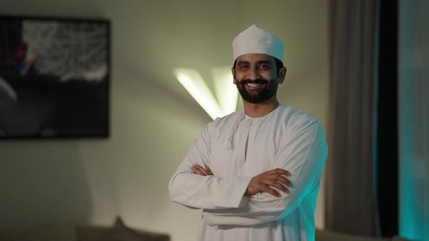 الوقوف بيدين متقاطعتين بإيماءات الثقة بالنفس، الاهتمام بالمظهر الخارجي، مفهوم الرجولة والوقار، بورتريه لرجل عربي خليجي عماني يرتدي الدشداشة التقليدية والكمه العمانية ينظر بايماءات السرور والسعادة