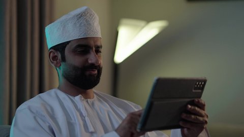 رجل عربي خليجي عماني يرتدي الدشداشة والكمة يحمل بيده جهاز التابلت فى غرفه المعيشة ، استخدام تطبيقات وبرامج الأجهزة اللوحية، الدردشة مع الأهل والأصدقاء، دمج التكنولوجيا والتقنيات الحديثة فى الحياه اليوميه ،تصفح مواقع التواصل الاجتماعي
