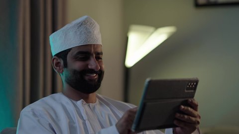 تصفح مواقع التواصل الاجتماعي، استخدام تطبيقات وبرامج الأجهزة اللوحية، الدردشة مع الأهل والأصدقاء، دمج التكنولوجيا والتقنيات الحديثة فى الحياه اليوميه ، رجل عربي خليجي عماني يرتدي الدشداشة والكمة يحمل بيده جهاز التابلت فى غرفه المعيشة