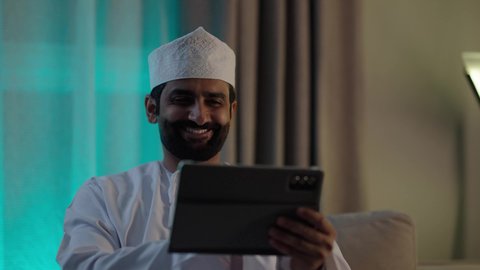 الدردشة مع الأهل والأصدقاء، استخدام تطبيقات وبرامج الأجهزة اللوحية، دمج التكنولوجيا والتقنيات الحديثة فى الحياه اليوميه ، رجل عربي خليجي عماني يرتدي الدشداشة والكمة يحمل بيده جهاز التابلت فى غرفه المعيشة ، تصفح مواقع التواصل الاجتماعي
