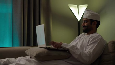 An Arab Gulf Omani man wearing a dishdasha and a kumma is working on a laptop with focused gestures in the living room, using advanced modern technologies, managing and organizing work affairs, and browsing social media.