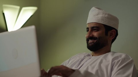 إدارة شؤون العمل وتنظيمه، تصفح مواقع التواصل الاجتماعي، رجل عربي خليجي عماني يرتدي الدشداشة والكمة يقوم بالعمل على الحاسوب المحمول بإيماءات التركيز فى غرفه المعيشة ، استخدام التقنيات الحديثة المتطورة