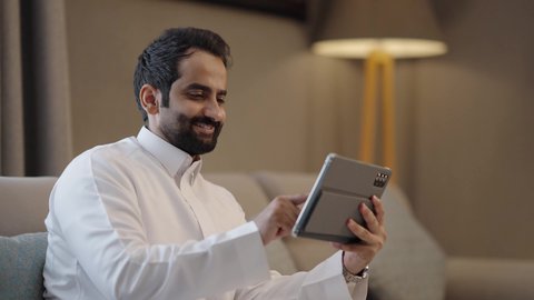 استعمال التكنولوجيا والتقنيات الحديثة، الدردشة مع الأهل والأصدقاء، رجل عربي خليجي سعودي يرتدي الثوب الابيض يجلس فى غرفه المعيشة يحمل بيده جهاز التابلت، تصفح مواقع التواصل الاجتماعي، استخدام تطبيقات وبرامج الأجهزة اللوحية،ايماءات السعادة و السرور