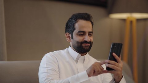 Exchanging views and ideas, browsing social media through a mobile phone, gestures of happiness and joy, an Arab Gulf Saudi woman wearing a hijab and abaya sitting in the living room with her husband holding a tablet.