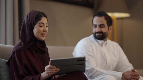 Integrating technology and tech into daily routines, discussing ideas and exchanging opinions, gestures of happiness and joy, an Arab Gulf Saudi woman wearing a hijab and abaya sitting in the living room with her husband holding a tablet.